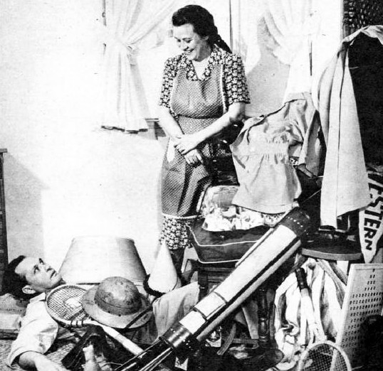 A black and white photograph of Fibber McGee and Molly, a couple from a popular radio show of the same name. They are standing in front of a closet that has been opened, with clothes and other items spilling out onto the floor. Fibber is holding a mop and wearing an apron, while Molly looks on with a smile on her face. The overall effect is one of humor and domesticity, with the couple's playful antics and cluttered living space providing a snapshot of life in mid-century America in 1948.