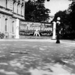 Buy Bonds: A large billboard on the exterior of a building promoting war savings stamps. It reads, "Pledge your quarters for victory, buy and invest wisely in War Savings Stamps." The text highlights that these are "guaranteed by U.S. Government” with "4% interest." The advertisement encourages supporting the war. The scene includes a street view with trees, a building with columns, and a decorative streetlamp in front.