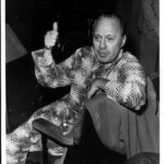 A black and white photograph of Jack Benny, a man in a suit and tie, standing against a plain background. He is looking off to the side with a slight smile on his face, and his hands are clasped in front of him. Benny was a comedian and actor, best known for his radio and television shows in the 1930s, 1940s, and 1950s. The photograph conveys a sense of classic Hollywood glamour and style, with Benny's sharp features and piercing blue eyes on prominent display.