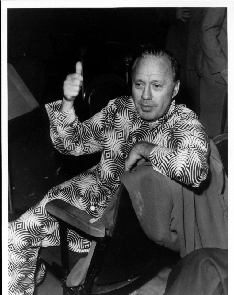 A black and white photograph of Jack Benny, a man in a suit and tie, standing against a plain background. He is looking off to the side with a slight smile on his face, and his hands are clasped in front of him. Benny was a comedian and actor, best known for his radio and television shows in the 1930s, 1940s, and 1950s. The photograph conveys a sense of classic Hollywood glamour and style, with Benny's sharp features and piercing blue eyes on prominent display.