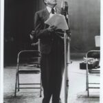 A black-and-white photograph depicting Jack Benny in a suit holding papers and speaking into a vintage-style microphone. The setting appears to be an old-time radio or recording studio. Jack is standing beside a couple of folding chairs, and there are some blurred background elements that suggest a studio environment. The overall tone is formal, reminiscent of mid-20th-century broadcasting.