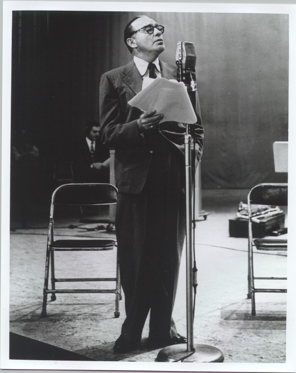 A black-and-white photograph depicting Jack Benny in a suit holding papers and speaking into a vintage-style microphone. The setting appears to be an old-time radio or recording studio. Jack is standing beside a couple of folding chairs, and there are some blurred background elements that suggest a studio environment. The overall tone is formal, reminiscent of mid-20th-century broadcasting.