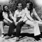 Lum, Abner and their real-life wives are sitting together on a bench outdoors, near a pool. They appear relaxed and are smiling at each other. The two people on the left are wearing casual attire, while the two on the right are dressed more formally, with the person on the far right in a light-colored suit and bow tie. Trees are visible in the background under a clear sky, suggesting a pleasant day.