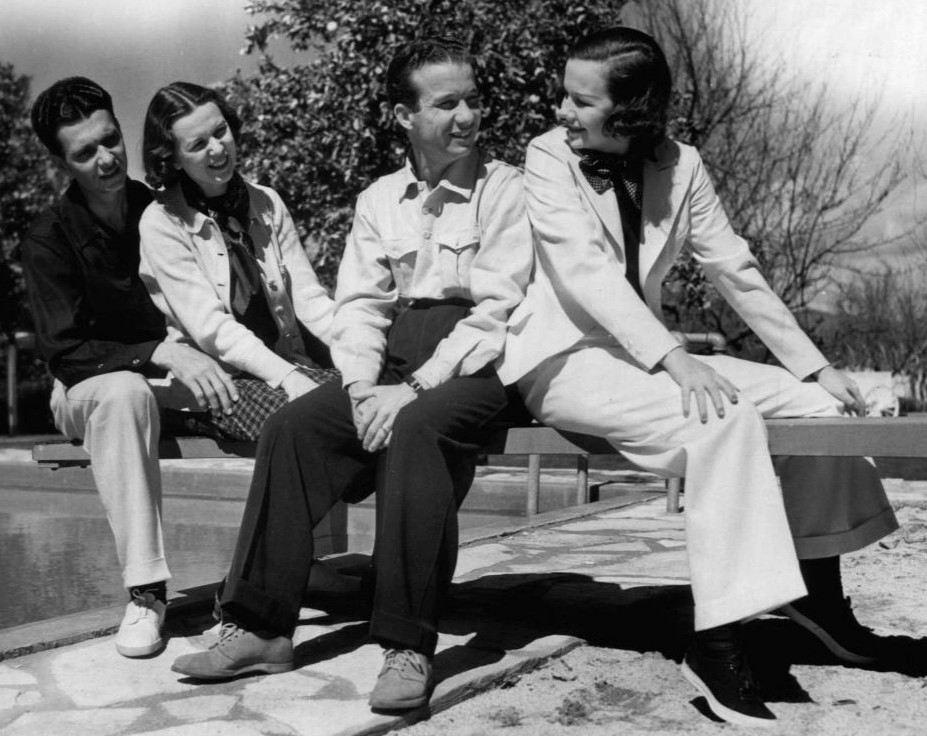 Lum, Abner and their real-life wives are sitting together on a bench outdoors, near a pool. They appear relaxed and are smiling at each other. The two people on the left are wearing casual attire, while the two on the right are dressed more formally, with the person on the far right in a light-colored suit and bow tie. Trees are visible in the background under a clear sky, suggesting a pleasant day.