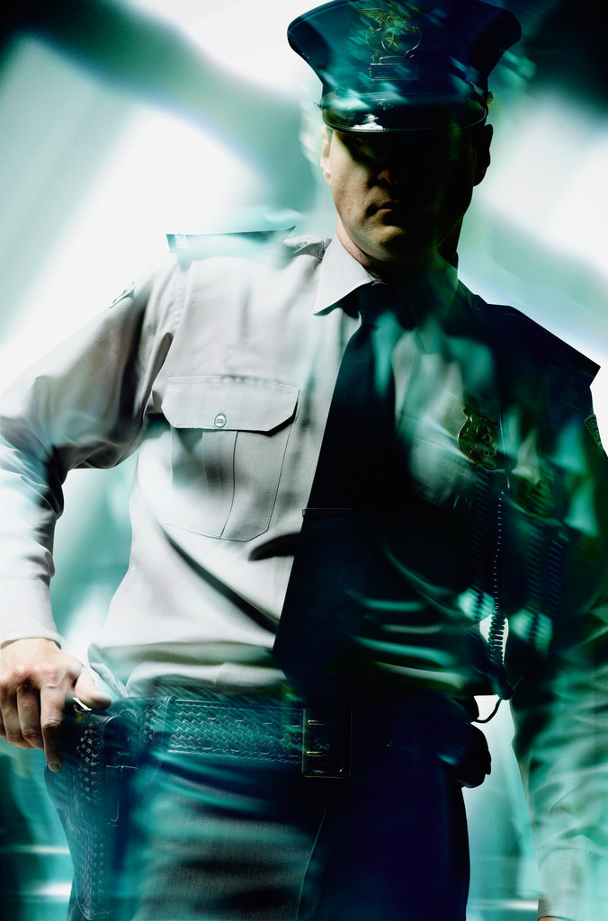 A security guard dressed in a dark uniform is reaching for his sidearm, which is holstered on his right hip. He stands in front of a brick wall, with his left hand resting on his belt. His facial expression suggests a state of high alertness and readiness to act in case of any threat or danger.