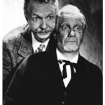 A black-and-white photo of Lum and Abner. Lum is on the left and has wavy hair and a mustache. He's wearing a suit and tie, and looks amused or curious. Abner is on the right, has messy hair and a beard, with exaggerated, surprised expression. He's wearing glasses and a dark suit with a bow tie. The composition suggests a comedic setting.