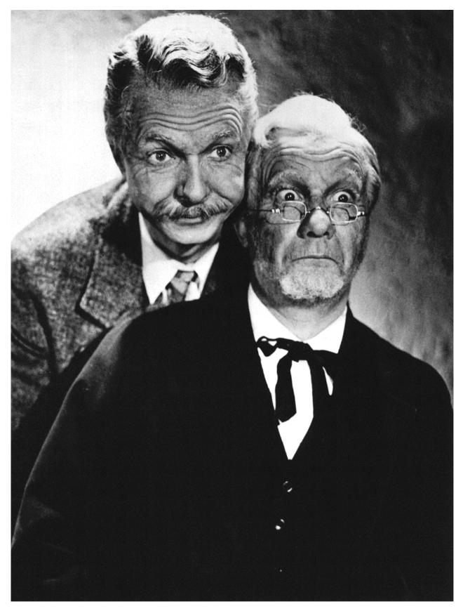 A black-and-white photo of Lum and Abner. Lum is on the left and has wavy hair and a mustache. He's wearing a suit and tie, and looks amused or curious. Abner is on the right, has messy hair and a beard, with exaggerated, surprised expression. He's wearing glasses and a dark suit with a bow tie. The composition suggests a comedic setting.