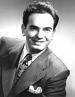 A black and white photograph of Elliott Lewis, a man in a suit and tie, standing in front of a microphone with headphones on. He appears to be speaking or reading from a script, with one hand raised to emphasize a point. The background is dark and nondescript, with no discernible details.