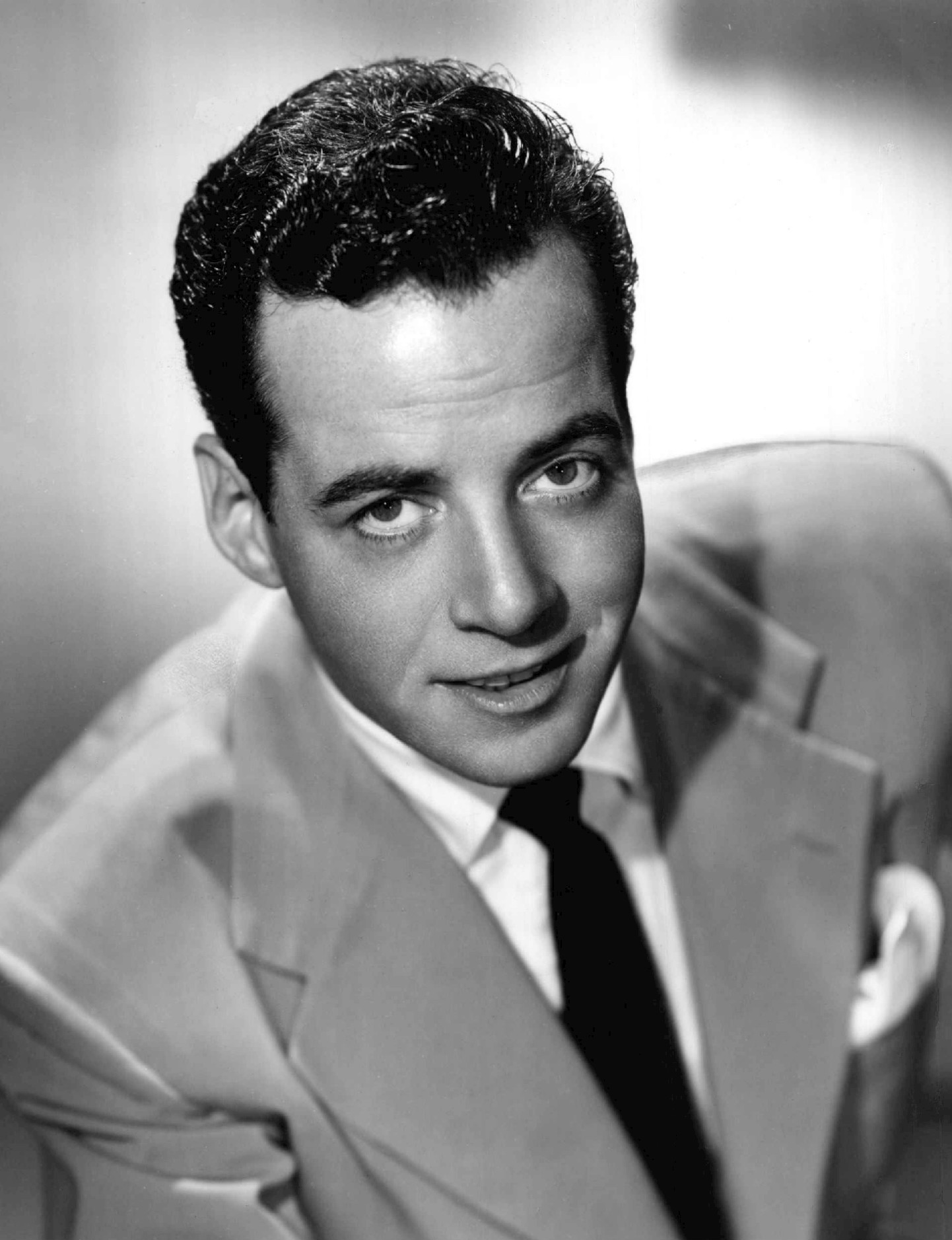 Gerald Mohr in 1951. A black and white photograph of Gerald Mohr, a man in a suit and tie, posing in front of a microphone with headphones on. He is holding a script in one hand and appears to be in the midst of speaking or recording. The background is dark and nondescript, with no discernible details.
