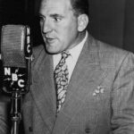 A black and white photograph of William Bendix as Chester Riley, a man in a suit and tie, standing at a microphone with headphones on. He is smiling and appears to be in the midst of speaking or recording. In the background, there is a sign that reads "Life of Riley," the name of the radio program he starred in. The overall effect is one of nostalgia and vintage Americana.