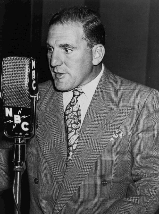 A black and white photograph of William Bendix as Chester Riley, a man in a suit and tie, standing at a microphone with headphones on. He is smiling and appears to be in the midst of speaking or recording. In the background, there is a sign that reads "Life of Riley," the name of the radio program he starred in. The overall effect is one of nostalgia and vintage Americana.