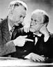 Lum and Abner in a Publicity Photo. 5x6. In this black and white publicity photo, two men, Lum and Abner, are posing together in a studio. They are dressed in suits and ties, with Abner on the left wearing a hat and holding a cane, while Lum on the right has his hands in his pockets. They are smiling at the camera with a friendly and approachable demeanor, creating a sense of warmth and familiarity. The photo has a vintage feel, reflecting the era in which they were popular radio personalities.