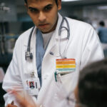 A photograph of a doctor wearing a stethoscope around their neck, standing against a plain background. The doctor is wearing a white lab coat and a serious expression on their face, as if listening intently to something. The stethoscope is draped over the doctor's shoulders and resting on their chest, and their hands are folded in front of them. The overall effect is one of professionalism and medical expertise.