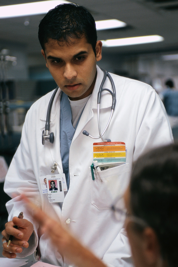 A photograph of a doctor wearing a stethoscope around their neck, standing against a plain background. The doctor is wearing a white lab coat and a serious expression on their face, as if listening intently to something. The stethoscope is draped over the doctor's shoulders and resting on their chest, and their hands are folded in front of them. The overall effect is one of professionalism and medical expertise.
