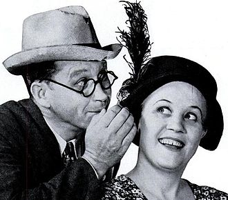 Jim Jordan in a Fedora and Marian Jordan wearing a hat in 1937. Standing in front of a microphone, dressed in vintage clothing from the 1930s, likely performing a radio show - the popular duo Fibber McGee and Molly.