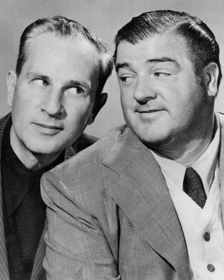 A black and white photograph of Abbott and Costello posing together. Abbott is on the left and has a slicked-back hairstyle and is looking to his right with a slight smile, while Costello on the right has a rounder face and is looking toward Abbott with a subtle grin. Both men are wearing plaid suits with shirts and ties. The background is plain, focusing attention on their expressions and attire.
