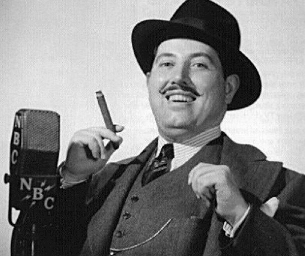 Harold Peary at the microphone, wearing a suit and hat. A black and white photograph of Harold Peary, a man in a suit and tie, smiling and looking off to the side. He is standing in front of a plain background, with his hands in his pockets. Peary was a comedian and actor, best known for his role as "The Great Gildersleeve" on radio and television in the 1940s and 1950s. The photograph conveys a sense of classic Hollywood glamour and style.