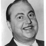 A close-up portrait of Willard Waterman as the Great Gildersleeves, a middle-aged man with slicked-back hair and a thin mustache. He is smiling widely, showing his teeth. He wears a dark suit jacket, white shirt, and patterned tie. The background is plain and light-colored, with no visible details. The lighting highlights the man’s facial features clearly, emphasizing his expressive eyes and cheerful demeanor. The overall look is formal yet friendly.