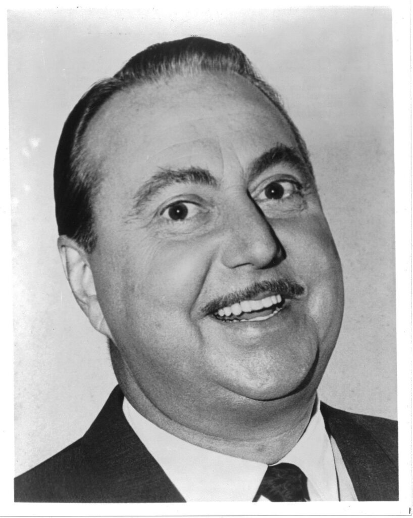 A close-up portrait of Willard Waterman as the Great Gildersleeves, a middle-aged man with slicked-back hair and a thin mustache. He is smiling widely, showing his teeth. He wears a dark suit jacket, white shirt, and patterned tie. The background is plain and light-colored, with no visible details. The lighting highlights the man’s facial features clearly, emphasizing his expressive eyes and cheerful demeanor. The overall look is formal yet friendly.